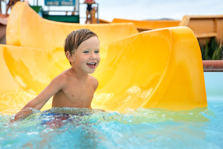 Cheerful blue-eyed kid on a water slide in the water park, a little boy merrily slides into the water along the slide in the amusement park, rest with children in the water park.