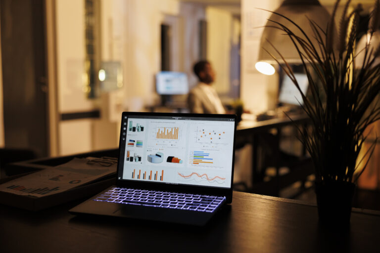 Laptop computer with marketing data report standing on table in startup office, in background employee working overtime at company strategy, brainstorming ideas for project. Business concept