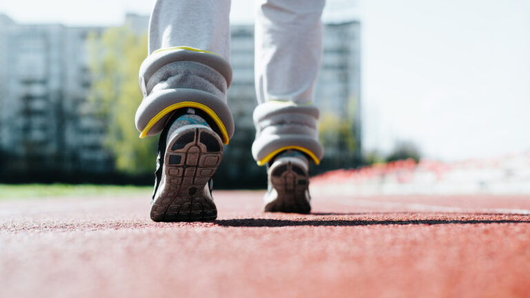 Low angle view of woman exercising in stadium outdoors. Close-up of an athlete with fitness weights walking on treadmill, back view. Selective focus on leg.