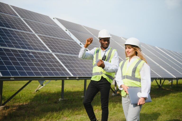 Multiracial people having working discussion about green energy while standing outdoors. Inspectors and foreman holding tablet, clipboard and drawings in hands. Solar panels arranged around.