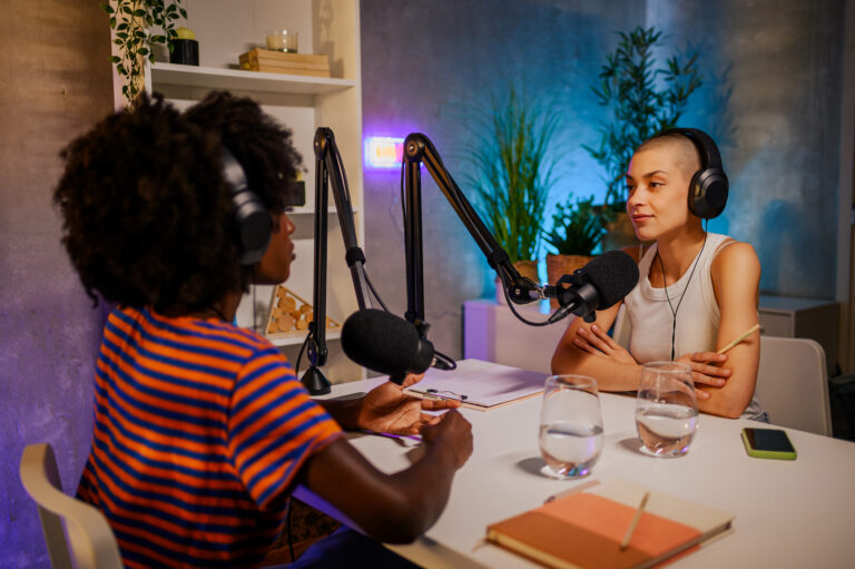 A young gen z girl is sitting at the table in a home broadcasting studio with headphones on and interviewing a multicultural guest while going live on air and live streaming for social media.